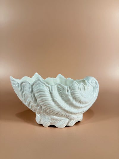 A white ceramic wall-mounted shell-shaped sconce with a sculpted seashell design, shown against a warm beige wall.