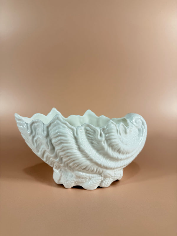 A white ceramic wall-mounted shell-shaped sconce with a sculpted seashell design, shown against a warm beige wall.