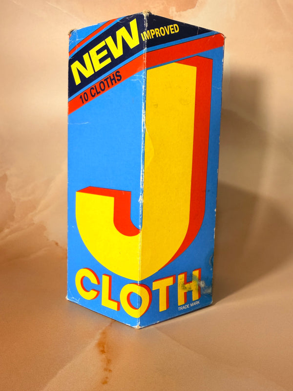 A vintage J Cloth box, featuring a bright blue background with a large yellow 'J' and red and yellow text, photographed on a beige surface.