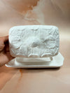 A hand holding a white ceramic butter dish with a lid featuring embossed leaf patterns, set against a warm beige marble background.