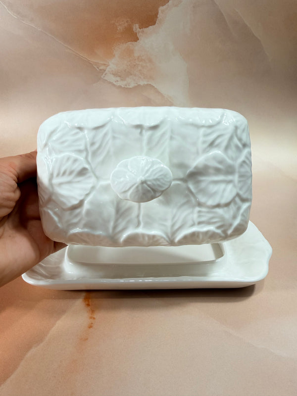 A hand holding a white ceramic butter dish with a lid featuring embossed leaf patterns, set against a warm beige marble background.
