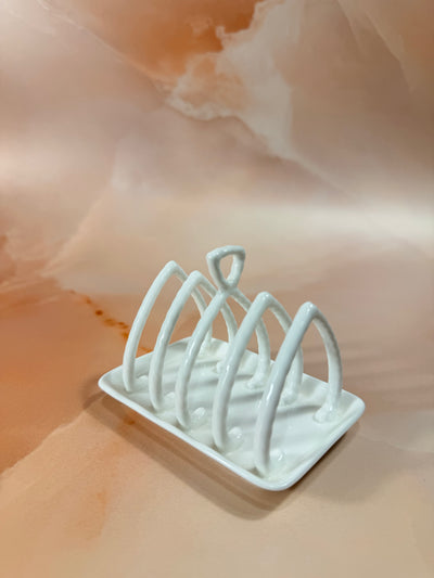 A white ceramic 5-slot bread toasting rack with a rectangular base and five upright loops, shown on a peach-colored marble surface.