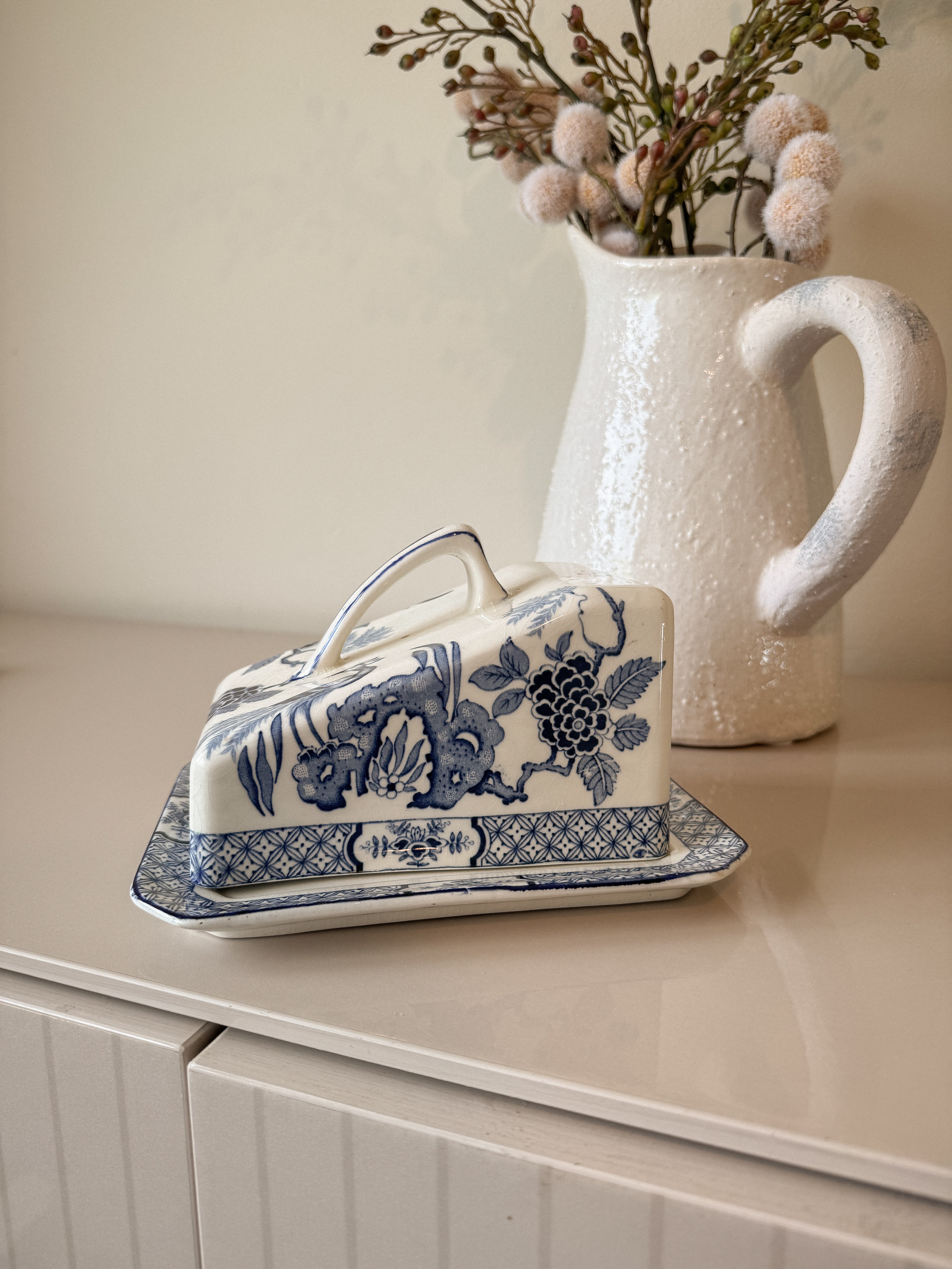 Decorative butter dish with floral design on a surface next to a vase with dried flowers.