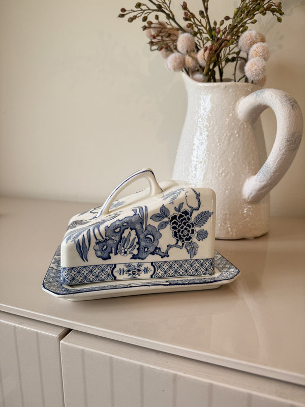 Decorative butter dish with floral design on a surface next to a vase with dried flowers.