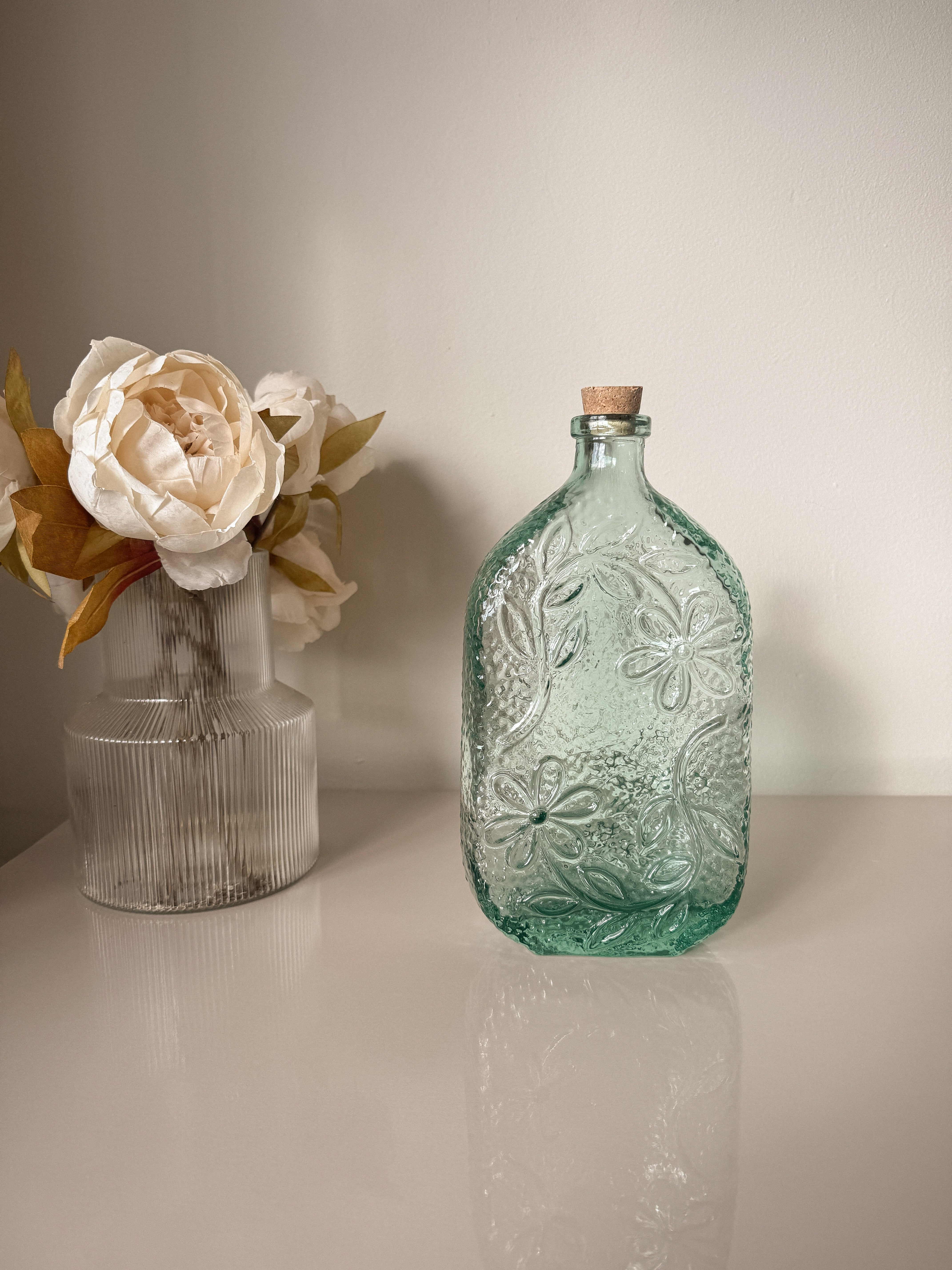 Decorative glass bottle with floral design next to a clear vase with flowers on a light surface.