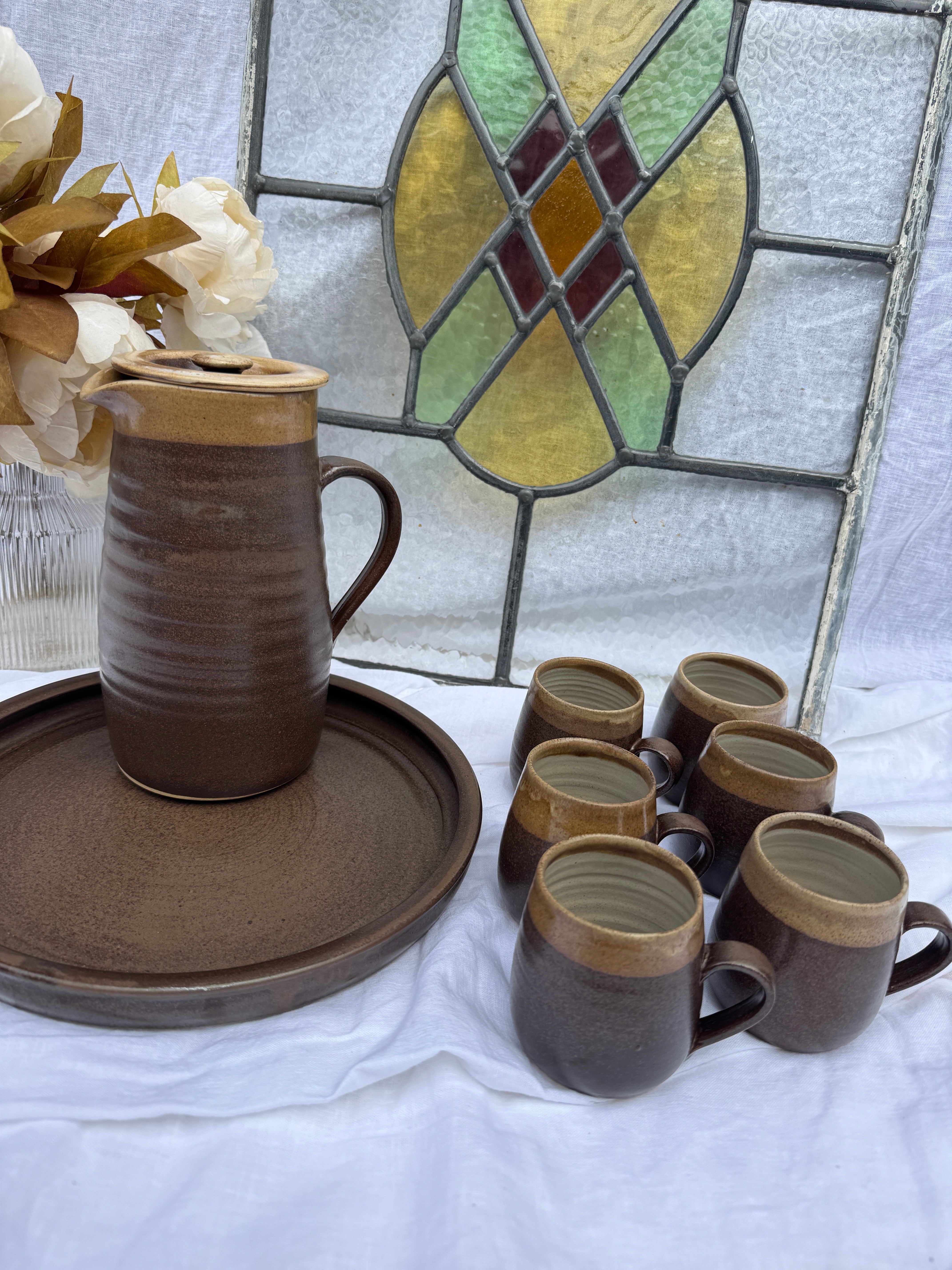 Set of ceramic mugs and a pot on a white surface with a stained glass window in the background.